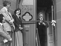 Man cutting a ribbon at an opening ceremony. To the left, a woman and man are standing and smiling 