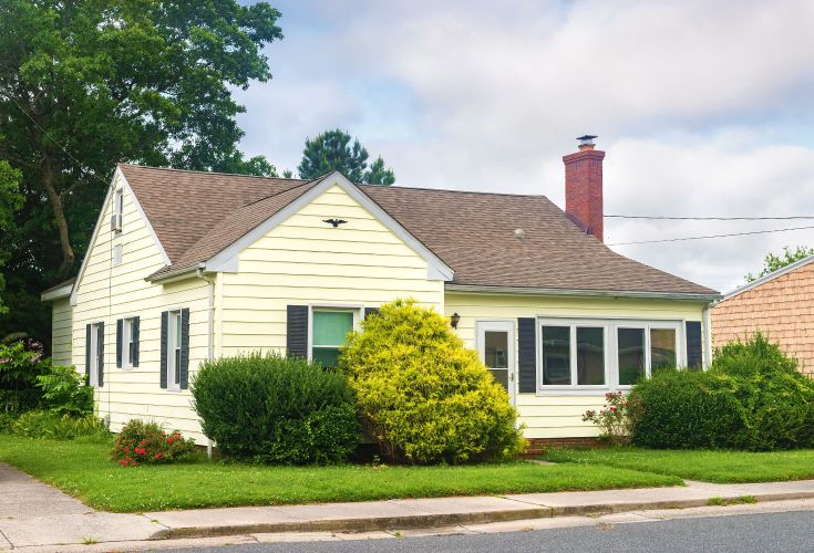 Front of a small yellow house with brown roof. The yard has large bushes and flowers
