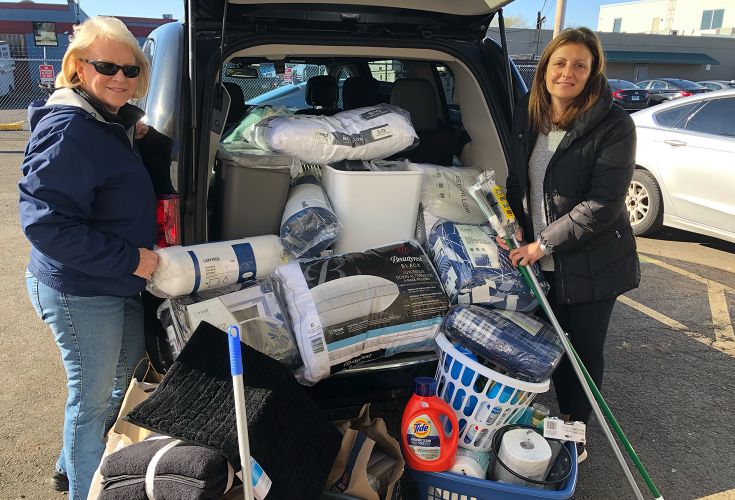 Two women standing in front of a car trunk that is overflowing with donated household items, such as pillows, detergent, and cleaning supplies
