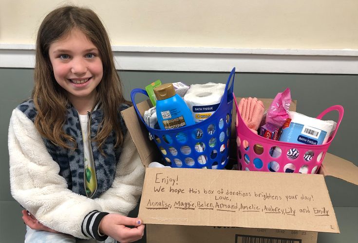 Young girl smiling next to a donation box full of different household items. The box has a note that reads "Enjoy! We hope this box o donations brightens your day! Love, Annalise, Maggie, Belen, Armand, Amelie, Aubrey, Lily, and Emily"