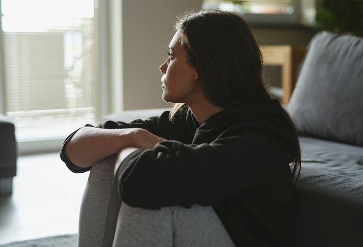 Girl sitting on the floor with her knees up against her chest, looking out the windw
