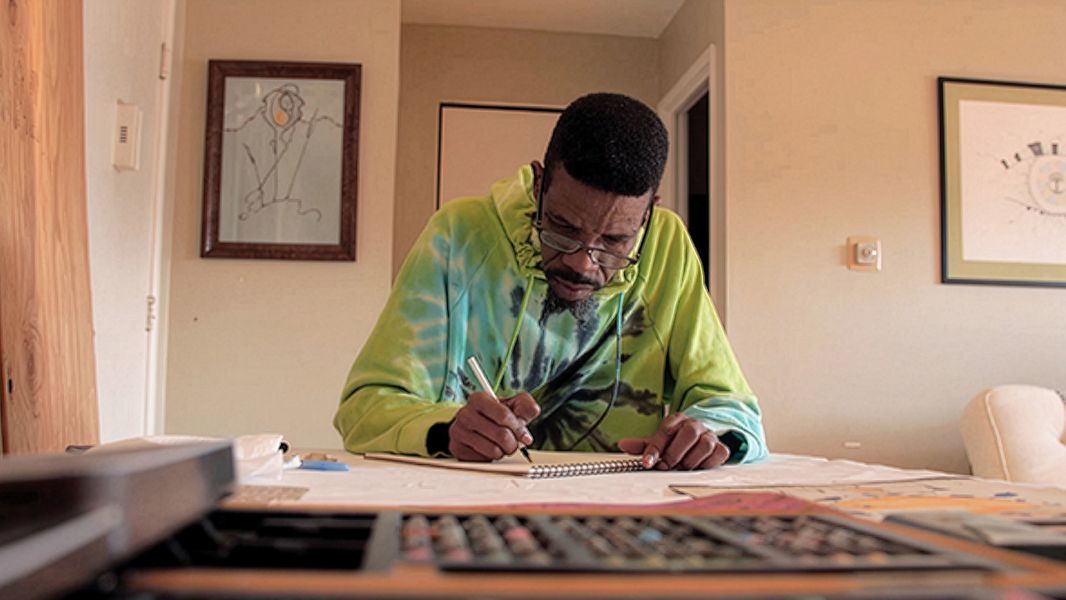 Man drawing on paper inside apartment with art supplies on table.