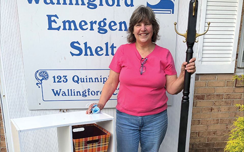 Pauline posing with furniture items in front of a sign that says "Wallingford Emergency Shelter"